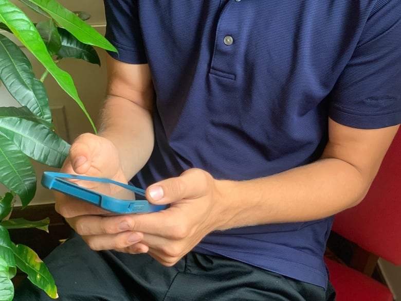 A young man's body and hands are seen scrolling through his phone while sitting in a bright pink chair next to a houseplant.